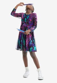 Blumendruckkleid mit lebendigen lila und türkisfarbenen Tönen, langen Ärmeln und einem ausgestellten Rock. Weiße Turnschuhe mit leicht strukturierten Socken.