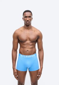 Μυώδης άντρας που στέκεται αντικρίζοντας μπροστά, φορώντας ανοιχτό μπλε μποξεράκια με τη ζώνη μέσης "HILFIGER", σε απλό ανοιχτό γκρι φόντο.