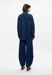 Διχτυωτό πουκάμισο με V-shaped yoke, μακριά μανίκια, κουμπωτές μανσέτες και χαλαρή εφαρμογή; συνδυασμένο με μπλε τζιν παντελόνι με ευρύ πόδι.