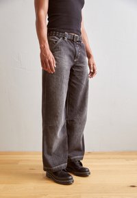 Svarta jeans med vida ben och hög midja, prydda med silverfärgade öglor, kontrasterande sömmar och en avslappnad passform, tillsammans med svarta stövlar.