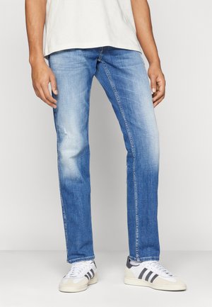 Jeans de mezclilla azul claro con un diseño de pierna recta, secciones deslavadas y un ligero efecto de arrugado, combinados con zapatillas blancas y grises.