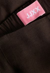 Nahaufnahme eines dunkelbraunen Stoffs mit gerafftem Bund und einem rechteckigen pinkfarbenen Etikett mit weißem Schriftzug "JJXX".