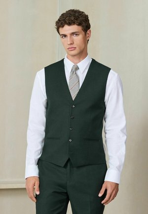 Young man with curly hair wearing a dark green waistcoat and matching trousers over a white shirt with a striped tie.