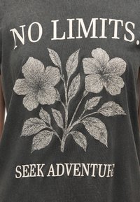 Σκούρο γκρι μπλουζάκι με λευκό κείμενο "NO LIMITS." πάνω και "SEEK ADVENTURE" κάτω, λεπτομερής απεικόνιση δύο μεγάλων λουλουδιών με φύλλα.