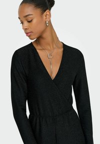 Vestido negro ajustado con mangas largas y un escote en pico profundo. La tela presenta un sutil brillo. Destacada cadena de plata con un colgante circular.