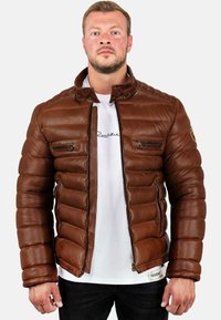 Bruine leren pufferjack met een opstaande kraag, ritszakken en een gewatteerde textuur. Draagt over een wit T-shirt en donkere spijkerbroek.