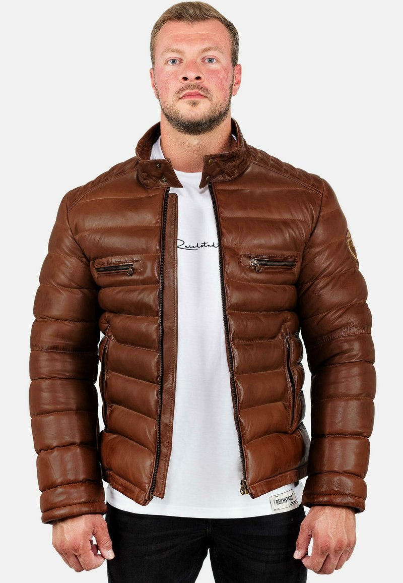 Bruine leren pufferjack met een opstaande kraag, ritszakken en een gewatteerde textuur. Draagt over een wit T-shirt en donkere spijkerbroek.