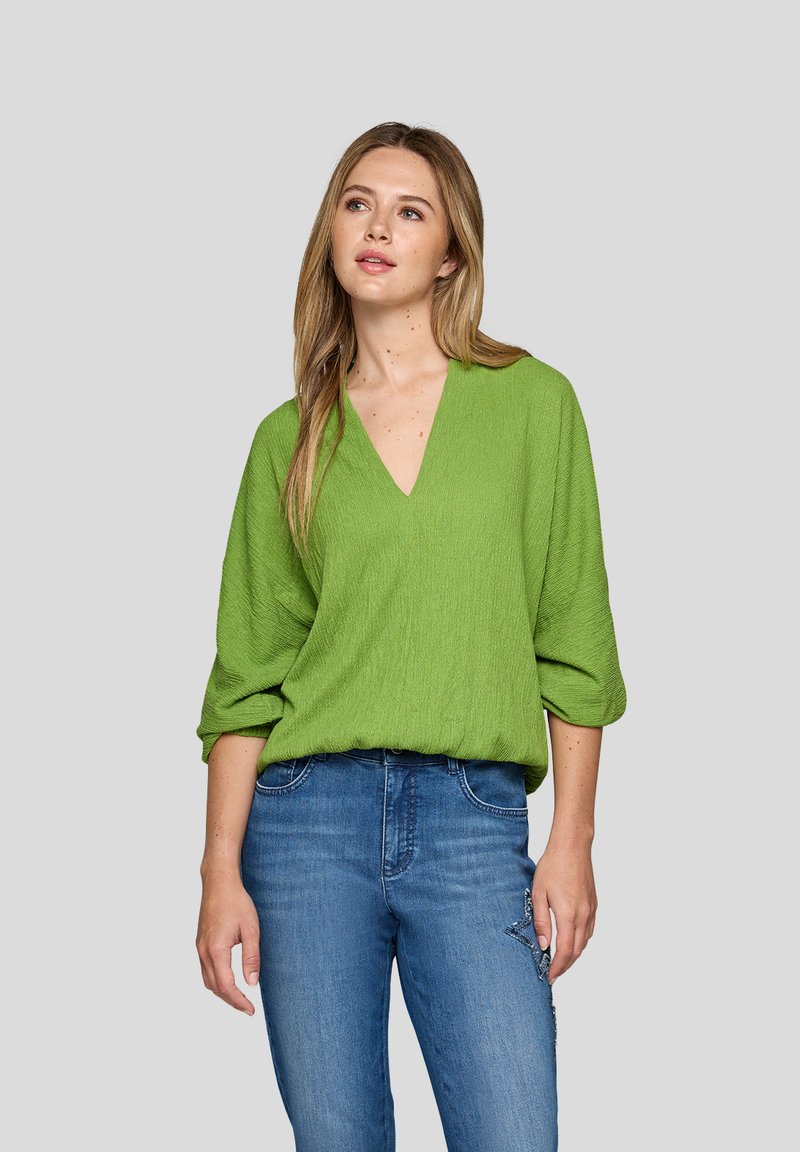 Frau mit langen blonden Haaren, die ein grünes V-Ausschnitt-Oberteil mit hochgerollten Ärmeln und eine blaue Jeans mit einem Stern-Patch trägt, vor einem neutralen Hintergrund.