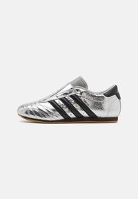 Hopeiset urheilukengät, joissa on teksturoitu metallipinta, mustat raidat ja harmaat yksityiskohdat, matala profiilimuotoilu, kumipohja.