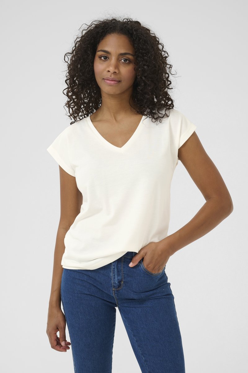 T-shirt crème à col en V en tissu doux avec des manches courtes retroussées. Associé à un jean fitted bleu foncé, mettant en valeur un look décontracté.