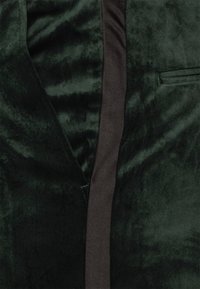Tessuto di velluto verde scuro con una trama liscia, caratterizzato da una striscia in raso marrone a contrasto e un dettaglio tasca sul lato sinistro.