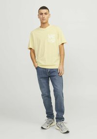 Gul kortærmet t-shirt med en hvid mønstret lomme, parret med blå jeans og grå træningssko, stående mod en neutral baggrund.