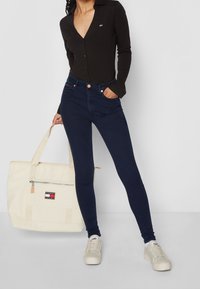 Camisa negra de botones, vaqueros ajustados azul oscuro, zapatillas crema y un bolso de mano crema con un logo rojo, blanco y azul.