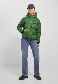 Grön puffjacka med huva, dragkedjestängning, kviltad design och framfickor. Matcher med blå jeans och svarta platåstövlar.