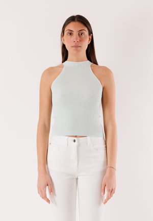 Donna con capelli lisci e scuri, che indossa un top senza maniche grigio chiaro a coste e pantaloni bianchi a vita alta, in piedi davanti a uno sfondo semplice.