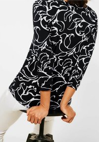 Person som bär en långärmad tröja med svart och vit mönster, sitter på en svart pall och greppar kanterna med båda händerna.