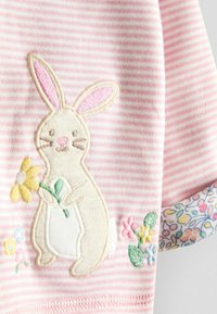 Ružovo-bielo pruhovaná látka s vyšívaným béžovým zajacom držiacim žltý kvet, obklopeným malými vyšívanými farebnými kvetmi.