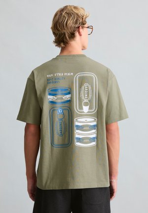 Persoon met kort blond haar en bril draagt een olijfgroen T-shirt bedrukt met blauwe en witte sardineblikillustraties en tekst op de achterkant.