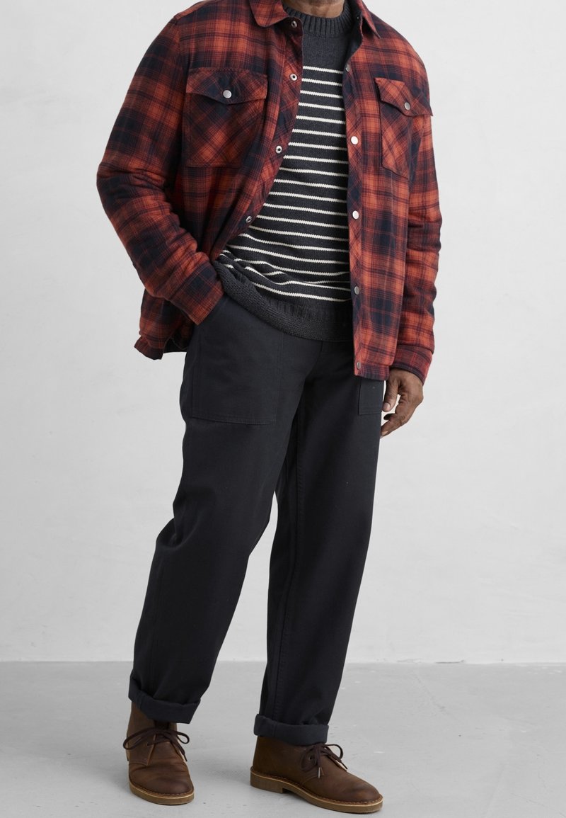 Homme portant une chemise à carreaux rouge et noire boutonnée par-dessus un pull à rayures noir et blanc, un pantalon noir et des chaussures marron à lacets.