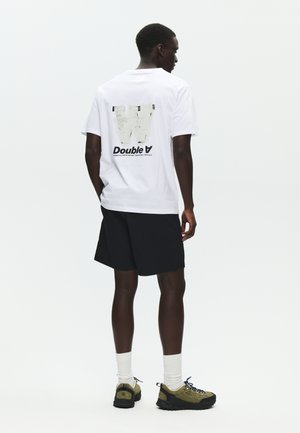 Mężczyzna stojący tyłem, ubrany w białą koszulkę z dużym wyblakłym napisem "W" i "Double A", czarne szorty, białe skarpetki i oliwkowo-zielone buty.