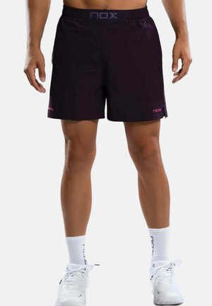Mann trägt schwarze Sportshorts mit "nox"-Logo, weiße Socken und weiße Sportschuhe, steht vor einem schlichten hellen Hintergrund.