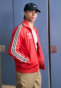 Red Adidas track jacket with black and white stripes on sleeves, standing pockets, smooth texture, paired with a black cap and beige pants.