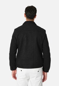 Giacca nera in misto lana con collo strutturato, polsini con bottoni e una texture liscia, abbinata a pantaloni bianchi. Vista posteriore mostrata.