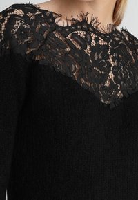 Pull noir avec un corps en tricot texturé et un panneau en dentelle transparente au niveau du col, présentant des motifs floraux et des bords effilochés le long de la dentelle.