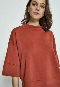 Rustfarvet oversized top med høj hals og korte faldne ærmer. Fremstillet af glat materiale med en afslappet pasform.