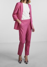 Růžové sako na míru a kalhoty ve stejném odstínu, doplněné bílým tričkem. Boty jsou tmavě fialové s ozdobnými kovovými detaily.