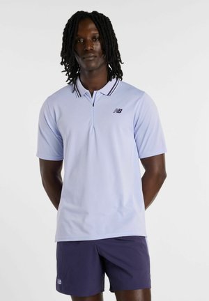 Mand med skulderlange dreadlocks iført lys lilla New Balance polo shirt og mørkelilla shorts, stående mod en ensfarvet baggrund.