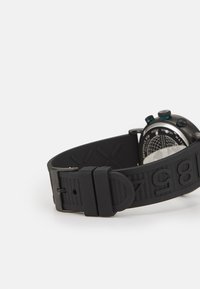 Correa de reloj de goma negra con números en relieve, hebilla ajustable y una esfera metálica con varios botones y un reverso texturizado.