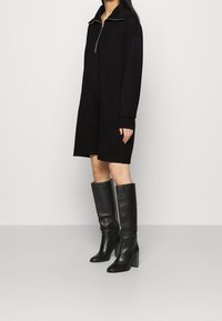 Mustat college-nilkkapituinen mekko, jossa on vetoketjukaulus, pitkät hihat ja rento malli. Yhdistetty polven yli ulottuviin mustiin nahkasaapikkaisiin, joissa on tukevat korot.