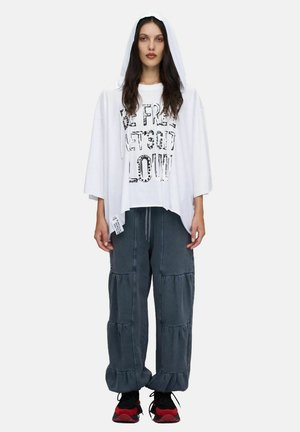 Femme portant une cape blanche à capuche, un t-shirt blanc imprimé, un pantalon large gris foncé avec des volants en étage, et des baskets noires et rouges, debout devant un fond blanc.