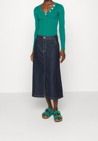 Mujer vestida con una blusa verde ajustada de manga larga y botones dorados, una falda midi de mezclilla oscura con una abertura en el frente y sandalias teal de doble tira con hebillas.