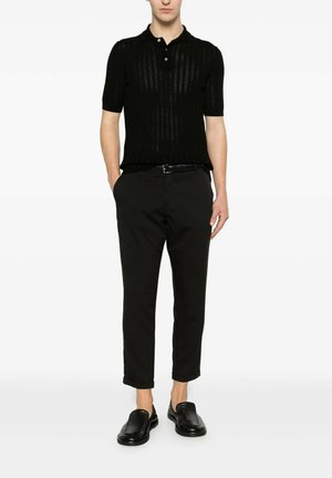 Uomo che indossa una polo a maniche corte in maglia nera, pantaloni neri corti con cintura e mocassini neri in pelle, in piedi con le mani in tasca.