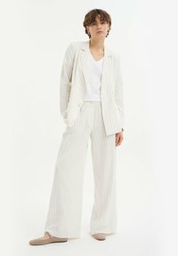 Completo a due pezzi di un crema chiaro con righe verticali. La giacca ha revers a faccetta e doppi bottoni; pantaloni a gamba larga e una camicia bianca a V.