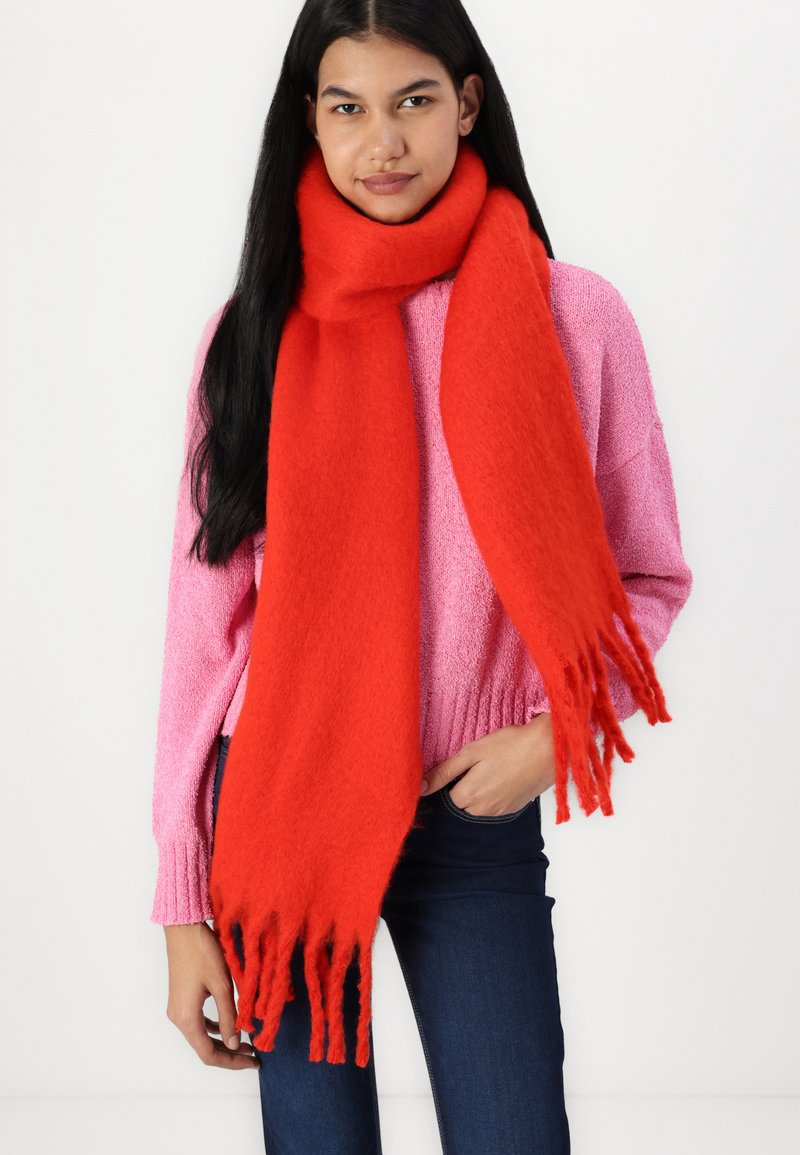Roter Schal aus weichem Material mit Fransen. Das Model trägt einen pinken, strukturierten Pullover und dunkle Jeans.