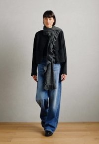 Zwarte cropped jack met zakken, gecombineerd met wijde blauwe jeans. Grijze gerimpelde sjaal voegt textuur toe. Model staat tegen een neutrale achtergrond.
