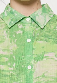 Chemise verte à motif avec col et boutons blancs, fabriquée en tissu léger texturé avec un design abstrait.