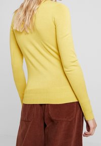 Žlutý pletený svetr s dlouhými rukávy, žebrovaným lemem a manžetami, přiléhavý střih. Kombinován s hnědými manšestrovými kalhotami, zdůrazňující kontrast textur.