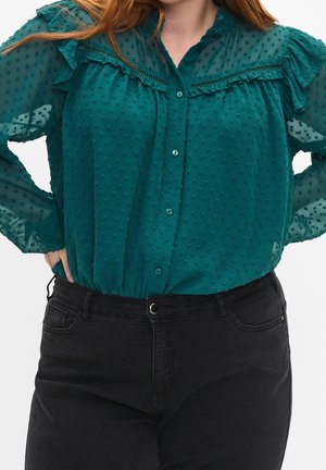 Femme portant un chemisier transparent texturé bleu sarcelle avec des volants et un jean noir taille haute, mains sur les hanches.