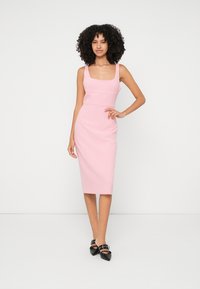 Ružičasta haljina prilagođenog kroja sa kvadratnim izrezom, dužine do koljena, glatke teksture i modernog kroja. Kombinirana s crnim cipelama na petu.