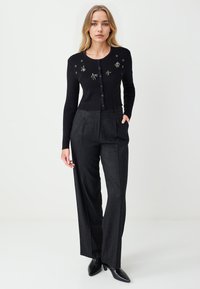 Cardigan noir côtelé avec broderie florale, associé à un pantalon gris taille haute et des escarpins noirs pointus. Élégance simple dans le design.