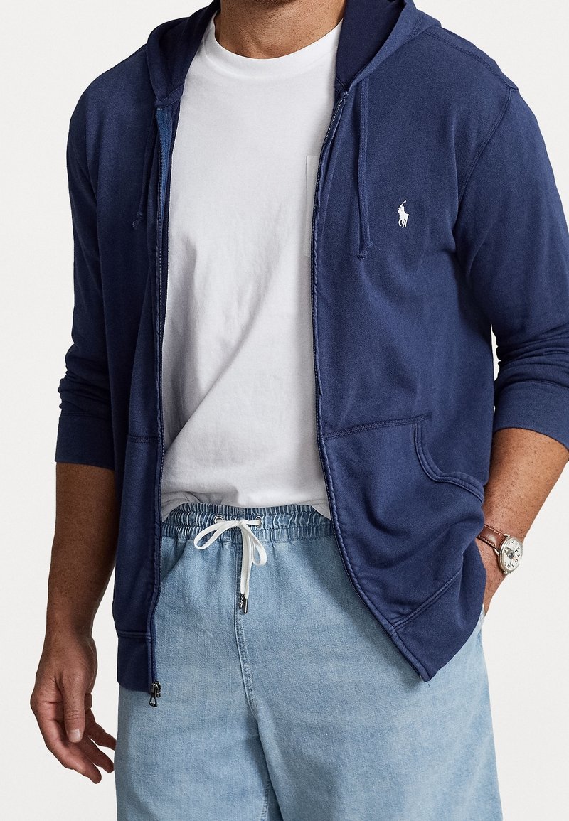 Marinblå zip-hoodie med framficka, bärs över en vit t-shirt med rund halsringning, kombinerat med ljusblå shorts med dragsko.