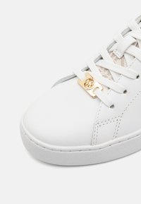 Weiße Ledersneaker mit einer strukturierten Gummisohle, ausgestattet mit goldenen Logo-Elementen und weißen Schnürsenkeln über einem atmungsaktiven Netzpaneel.