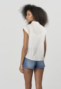 Fehér csíkos rövid ujjú blúz galléros nyakkivágással, kék farmer rövidnadrággal párosítva. A blúz könnyű, texturált anyagból készült.