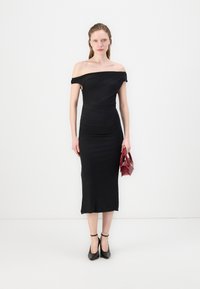 Svart off-the-shoulder kjole med rynket tekstur, kne-lengde. Modellen holder en bordeaux håndveske, kombinert med svarte ankelhæler.