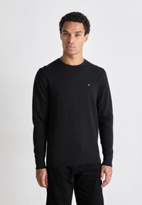 Maglione nero a maniche lunghe con tessuto testurizzato, scollo rotondo e piccolo logo bianco sul petto. Abbinato a pantaloni neri.