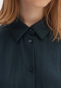 Camicia verde scuro con una tessitura strutturata, dotata di colletto, quattro bottoni e righe verticali. Vista ravvicinata che mette in evidenza il scollo.